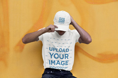 Tee Mockup Featuring a Man with a Dad Hat Leaning Against a Yellow Wall 